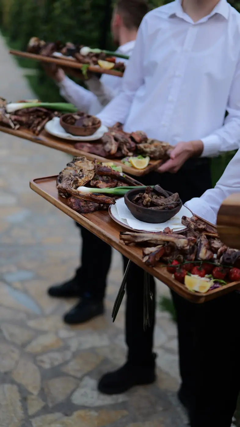 Waiters carrying long boards of grilled specialties through the garden