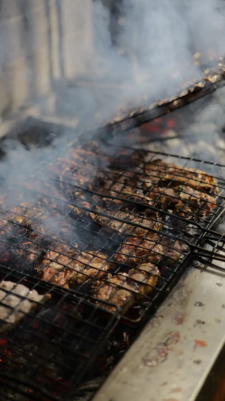Meat grilling over charcoal with smoke rising at OKO Farm